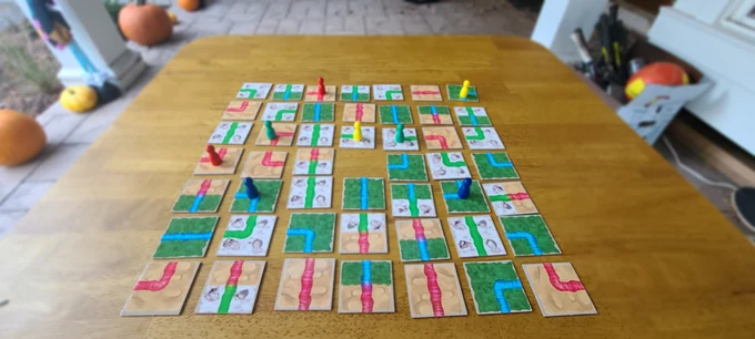 A Rainbow River game in progress on a wooden table: square tiles with red, blue and green rivers laid out in a grid, with colored player pawns